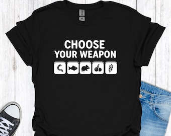 Op de afbeelding: Zwart T-shirt met de witte tekst "CHOOSE YOUR WEAPON". Daaronder vier witte iconen: een worm, een vis, een muis, kersen en een tarwe-aar. Het T-shirt wordt gedragen met een blauwe spijkerbroek en sneakers.