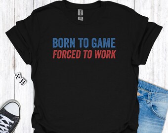 Op de afbeelding: Zwart T-shirt met de tekst "BORN TO GAME FORCED TO WORK" in blauw en rood. Het shirt wordt gecombineerd met een blauwe spijkerbroek en een paar dobbelstenen. Een paar zwart-witte sneakers is links onderin zichtbaar.