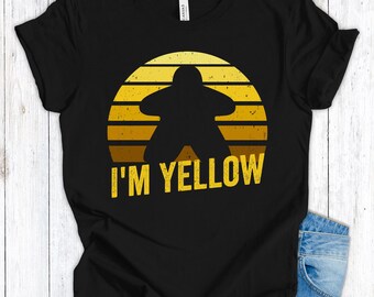 Op de afbeelding: Zwart T-shirt met een gele afbeelding van een bordspelstuk voor een gele zonsondergang. De tekst "I'M YELLOW" is in het geel onder de afbeelding gedrukt. Het shirt is gemaakt van zacht materiaal.