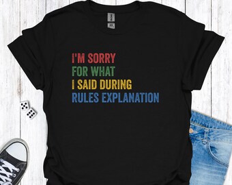 Könnte beinhalten: Schwarzes T-Shirt mit dem Text "I'M SORRY FOR WHAT I SAID DURING RULES EXPLANATION" in Rot, Grün, Gelb und Blau. Das Shirt wird mit einer blauen Jeans und Turnschuhen kombiniert.