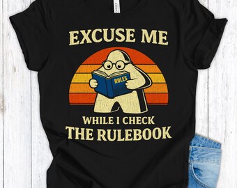 Op de afbeelding: Zwart T-shirt met een grafisch ontwerp. Het toont een stripfiguur met een bril die een boek leest met het label "RULES". De tekst hierboven luidt "EXCUSE ME" en hieronder "WHILE I CHECK THE RULEBOOK". Het ontwerp bevat een zonsondergang.