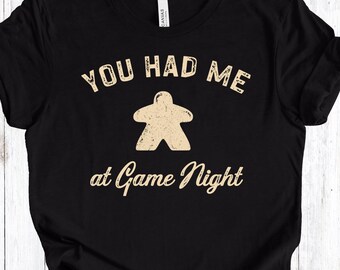 Op de afbeelding: Zwart T-shirt met de tekst "YOU HAD ME" boven een beige spelstukafbeelding en "at Game Night" eronder. Het shirt is gemaakt van een zacht materiaal en is geschikt voor vrijetijdskleding.