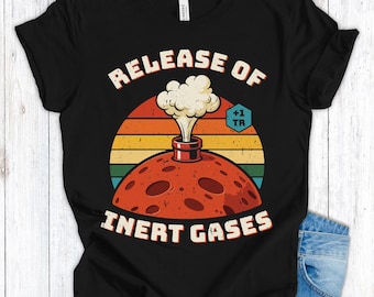 Op de afbeelding: Zwart T-shirt met een retro grafisch ontwerp. Het ontwerp toont een rode planeet met een rokende opening tegen een zonsondergang. De tekst luidt "RELEASE OF INERT GASES" in een vintage lettertype.