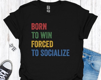 Op de afbeelding: Zwart t-shirt met de tekst "BORN TO WIN FORCED TO SOCIALIZE" in rood, groen, geel en blauw. De afbeelding bevat een blauwe spijkerbroek en een paar dobbelstenen.