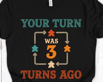 Op de afbeelding: Zwart t-shirt met de tekst "YOUR TURN WAS 3 TURNS AGO" in teal en oranje. Het ontwerp bevat spelstukpictogrammen en een kader met pijlen rond het nummer 3.