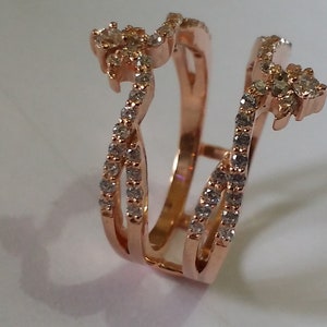 May include: Rose gold ring guard with a floral design at the top and sparkling stones. The ring features a double band design with a series of small, clear stones set along the bands. The ring is designed to be worn with another ring.