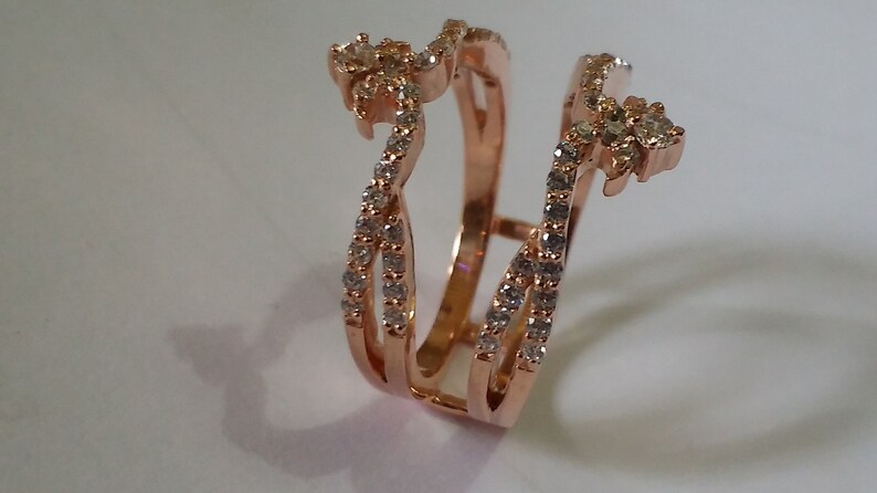 May include: Rose gold ring guard with a floral design at the top and sparkling stones. The ring features a double band design with a series of small, clear stones set along the bands. The ring is designed to be worn with another ring.
