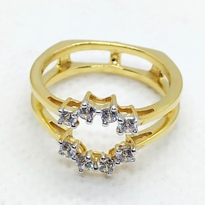 May include: A gold-toned ring with a double band design. The ring features a series of small, clear gemstones set in a curved pattern on the lower band and a similar arrangement on the upper band. The ring is set against a white background.