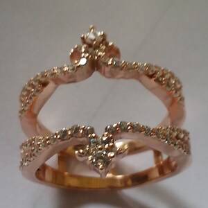 May include: Rose gold ring with a crown design, encrusted with small, clear gemstones. The ring features two bands, one forming the base and the other a crown shape, both adorned with sparkling stones. A small gemstone sits at the crown's peak.