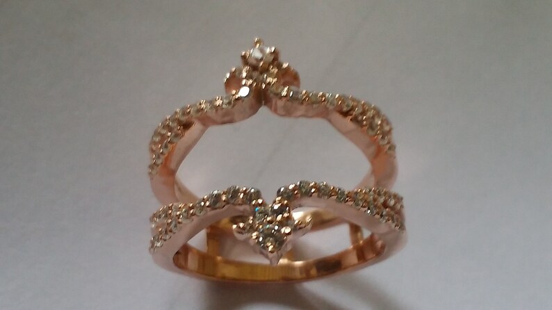 May include: Rose gold ring with a crown design, encrusted with small, clear gemstones. The ring features two bands, one forming the base and the other a crown shape, both adorned with sparkling stones. A small gemstone sits at the crown's peak.