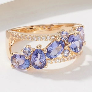 Puede incluir: Un anillo de oro con un diseño de doble banda que presenta un grupo de piedras preciosas moradas y transparentes. El anillo está engastado con piedras de varios tamaños y formas, incluyendo piedras en forma de pera y redondas.