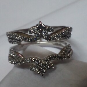 May include: A silver-toned ring with a crown-like design. The ring features a series of small, sparkling stones set along the band and in the crown's details. The ring is displayed on a white surface, showcasing its intricate design.