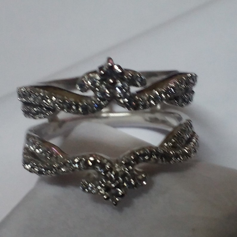 May include: A silver-toned ring with a crown-like design. The ring features a series of small, sparkling stones set along the band and in the crown's details. The ring is displayed on a white surface, showcasing its intricate design.