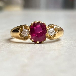 Könnte beinhalten: Ein goldener Ring mit einem großen roten Rubin-Edelstein und zwei kleineren Diamanten.