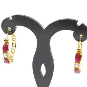 May include: Gold hoop earrings with red and white stones. The earrings are on a black display stand.