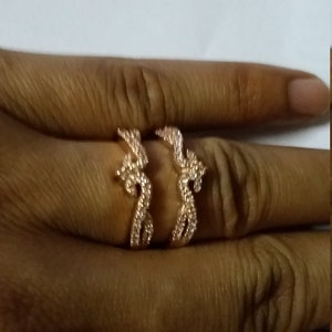 May include: A rose gold ring with a unique, open design. The ring features a swirling, abstract pattern, embellished with small, sparkling stones. The ring is shown on a finger, highlighting its intricate details and elegant style.