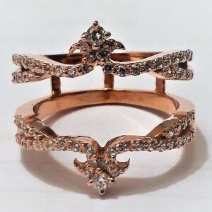 May include: Rose gold ring with a double band design. The ring features a crown-like top and bottom, both adorned with small, clear gemstones. The bands are also embellished with gemstones, creating a sparkling effect. The ring is set against a white background.