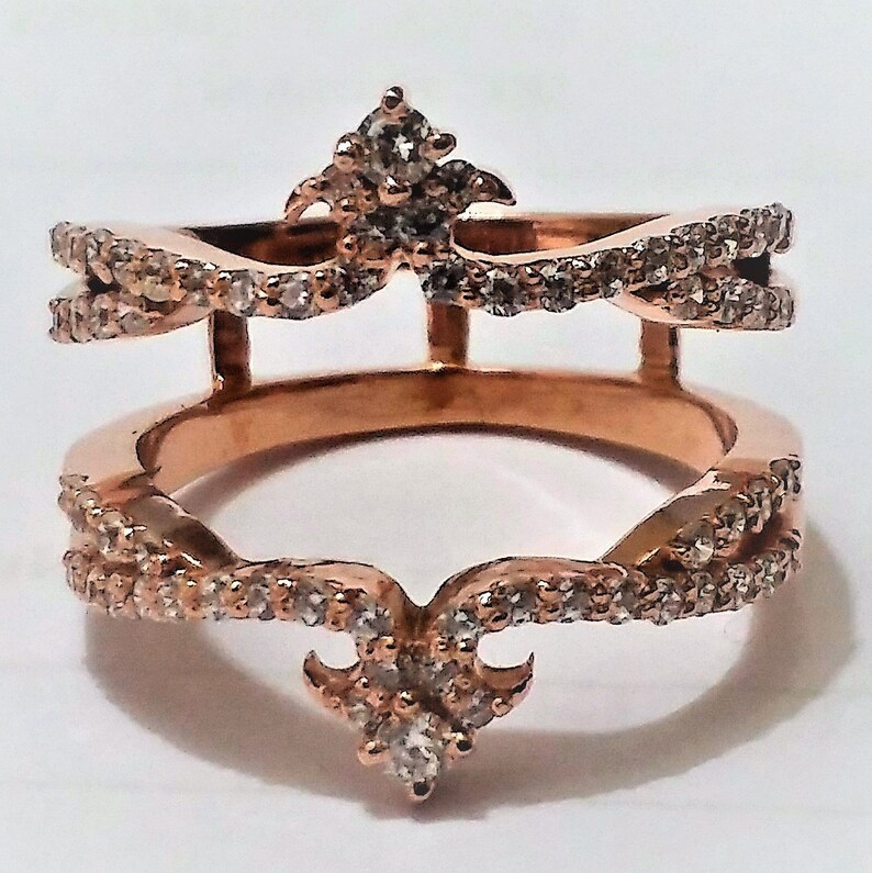 May include: Rose gold ring with a double band design. The ring features a crown-like top and bottom, both adorned with small, clear gemstones. The bands are also embellished with gemstones, creating a sparkling effect. The ring is set against a white background.