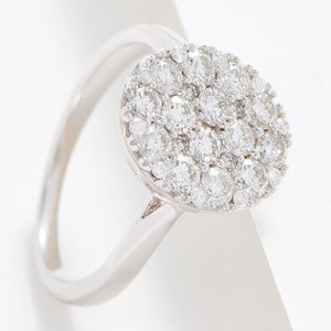 Puede incluir: Un anillo de plata con un grupo circular de pequeñas piedras preciosas claras. Las piedras están muy juntas, creando un efecto brillante. El anillo tiene una banda de plata pulida simple. El diseño es elegante y llamativo.