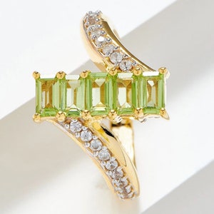 Puede incluir: Anillo de oro con una fila de cinco piedras preciosas verdes y una banda curva adornada con pequeñas piedras preciosas blancas.