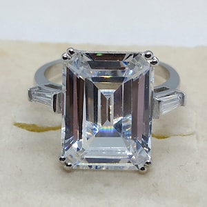 Puede incluir: Un anillo de plata con una gran piedra preciosa de talla esmeralda transparente y dos piedras preciosas más pequeñas de talla baguette a los lados.