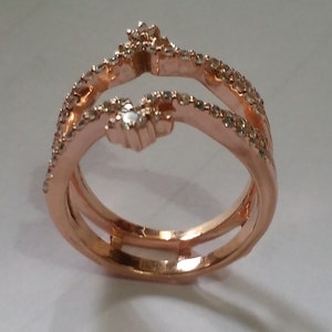 May include: Rose gold ring with a unique design. The ring features three bands, with two bands curving upwards and meeting at the top. The bands are adorned with small, sparkling stones. The ring is set against a white background.