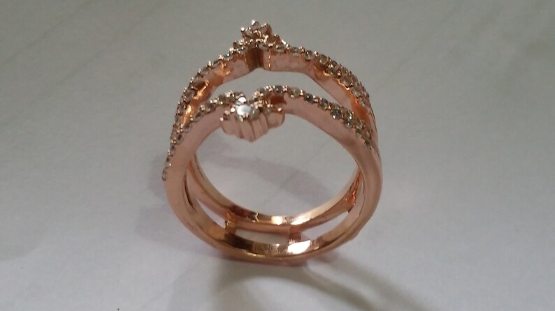May include: Rose gold ring with a unique design. The ring features three bands, with two bands curving upwards and meeting at the top. The bands are adorned with small, sparkling stones. The ring is set against a white background.