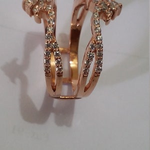 May include: Rose gold ring with a unique design. The ring features multiple bands adorned with small, clear gemstones. The design has an open center and decorative elements on the sides, creating a distinctive and elegant look.