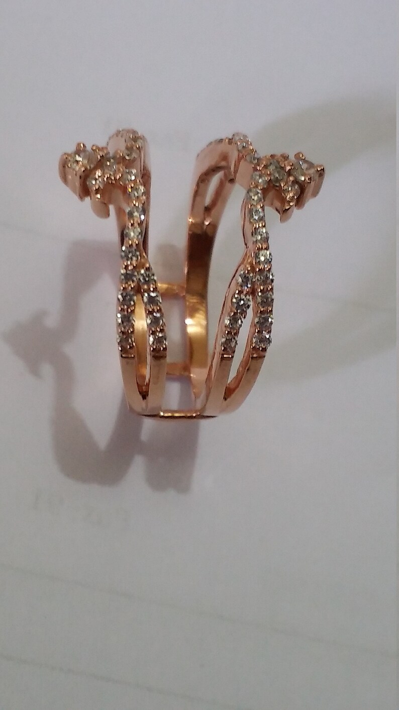 May include: Rose gold ring with a unique design. The ring features multiple bands adorned with small, clear gemstones. The design has an open center and decorative elements on the sides, creating a distinctive and elegant look.