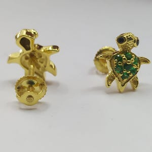May include: A pair of gold turtle stud earrings with green and black gemstones. The earrings are designed with a detailed turtle shape and feature a post back closure.