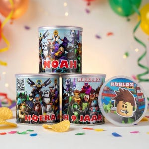 Op de afbeelding: Drie cilindrische containers met kleurrijke Roblox-personageontwerpen. Eén container heeft de naam "NOAH" erop gedrukt. De andere containers hebben de tekst "ROBLOX" en "HOERA! 9 JAAR". De containers staan op een oppervlak met confetti en chips.
