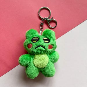 May include: Green plush frog keychain with a sad expression, red cheeks, and black eyes. The keychain is attached to a silver metal ring.