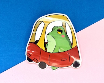 Peut inclure: Une grenouille verte conduisant une voiture jouet rouge avec un toit jaune. La grenouille a un œil jaune et un rouge sur la joue.