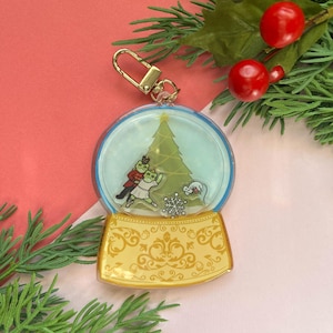 May include: A clear glass snow globe keychain with a gold base. The snow globe features a miniature Christmas tree, a couple dancing, and a snowflake. The base is decorated with a gold floral pattern.