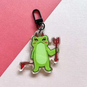 Könnte beinhalten: Grüner Frosch-Schlüsselanhänger mit roten Teufelshörnern und einem roten Dreizack. Der Frosch hat einen mürrischen Gesichtsausdruck und hält den Dreizack in seiner rechten Hand.