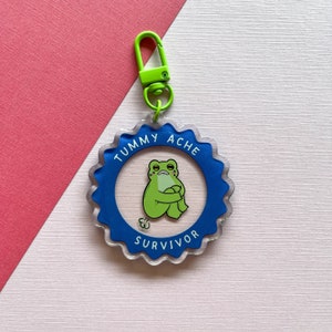 May include: A blue and clear acrylic keychain with a green frog cartoon illustration. The keychain has the text "Tummy Ache Survivor" around the edge.