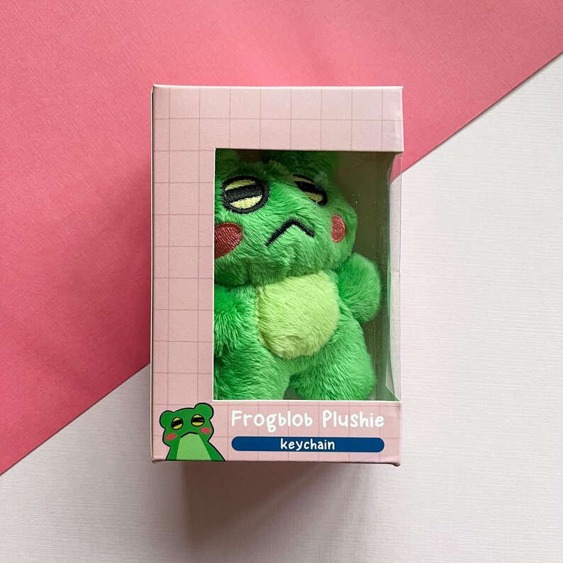 May include: A green frog plush keychain with a grumpy expression. The plush is packaged in a pink and white box with the text "Frogblob Plushie keychain".