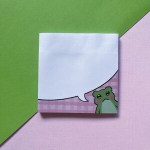 May include: A pink and white gingham notepad with a speech bubble and a green frog with yellow eyes. The frog has a pink blush on its cheeks.