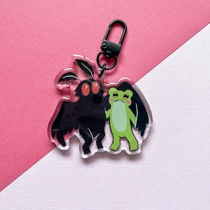 May include: A clear acrylic keychain featuring a black moth-like creature with red eyes and a green frog with black wings. The keychain has a black clasp and is set against a pink and white background.