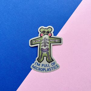 May include: A green sticker in the shape of a cartoon frog with a skeleton design inside. The sticker has the text "I'M FULL OF MICROPLASTICS" in blue and white. The background is blue and pink.