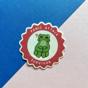 May include: A red and white sticker with a cartoon frog with big eyes and a worried expression. The sticker says "Panic Attack Survivor" in a white circle.