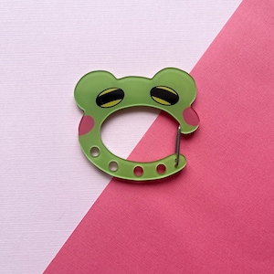 May include: A light green frog-shaped carabiner with a smooth finish. The design features two dark oval eyes with yellow accents, pink circular cheek patches, and four small circular cutouts along the bottom curve of the metal-hinged frame.