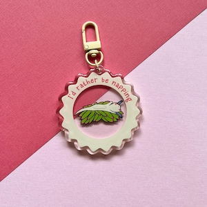 May include: A white and pink acrylic keychain with a gold metal ring. The keychain features a cartoon axolotl sleeping on a bed of green leaves. The text "I'd rather be napping" is written around the edge of the keychain.