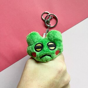 May include: Green plush keychain with a sad face, big eyes, and red cheeks. The keychain is attached to a silver key ring.