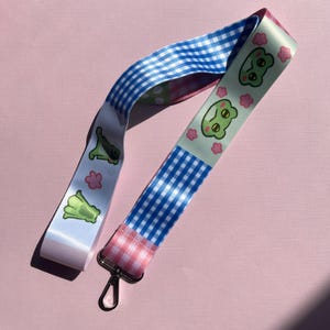 Könnte beinhalten: Ein Schlüsselband mit Metallverschluss. Es zeigt ein sich wiederholendes Muster aus blauem Vichy, rosa Vichy und einem weißen Band mit grünen Froschillustrationen und rosa Blumenakzenten. Das Schlüsselband befindet sich auf einem rosa Hintergrund.