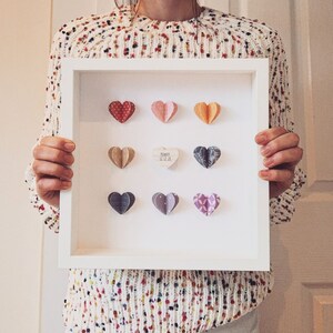 May include: White frame with a white mat and nine paper hearts. The hearts are in various colors and patterns. The center heart is a light brown color with the text "Married" and the date "12.12.21".