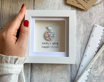 May include: A white frame with a map of a city folded into the shape of a ring. The text "Kirsty & Greg Engaged 23.11.2021" is written below the ring.
