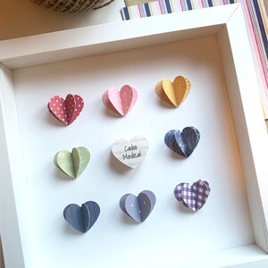 May include: A white frame with a white background containing nine paper hearts. The hearts are decorated with different patterns, including polka dots, plaid, and floral designs. The text "Cadoo Medical" is written on one of the hearts.