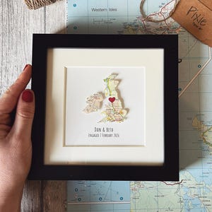 May include: A framed map of the UK, with a red heart marking a location. The frame is black. The text "DAN & BETH ENGAGED 7 FEBRUARY 2026" is below the map. A hand holds the frame. A tag with the word "Pixie" is visible.
