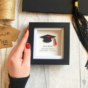 May include: A black shadow box frame with a white mount. Inside the frame is a black paper graduation cap with a red tassel. The text "JOANNE WRIGHTY OXFORD BROOKES UNIVERSITY BA HONS SOCIOLOGY - CLASS OF 2022" is printed in black on a white background.